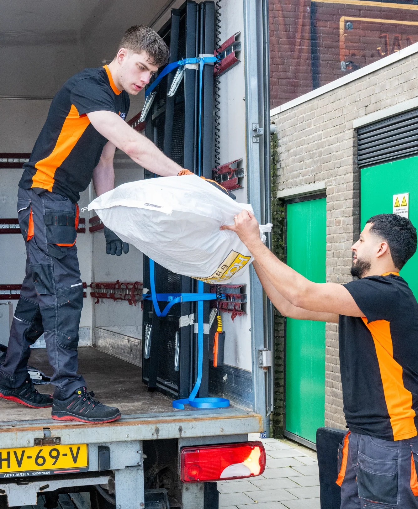 Twee medewerkers voeren een bedrijfsontruiming uit en laden grote zakken op een vrachtwagen met laadklep