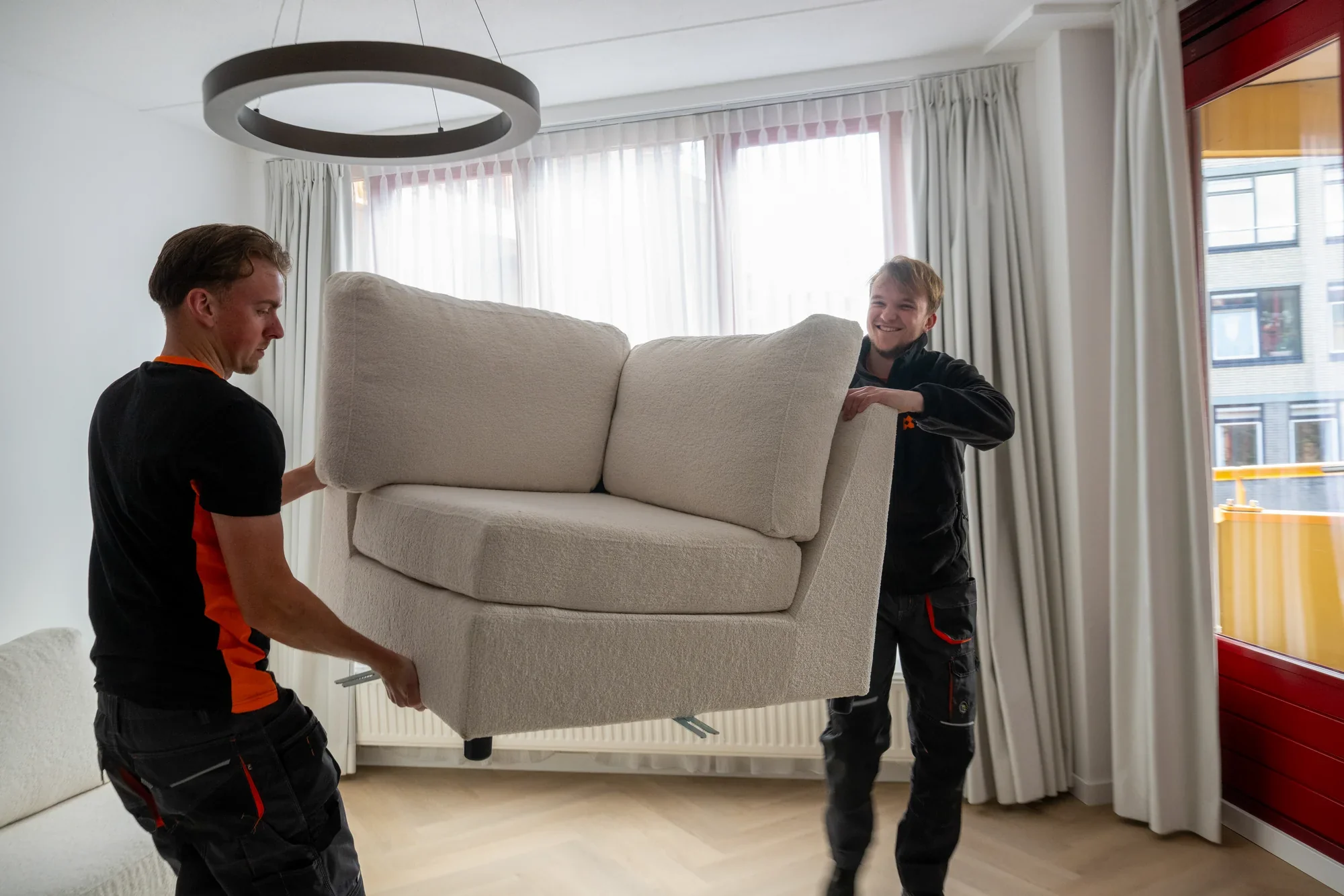 Two Nova staff lifting a sofa in a living room with curtains